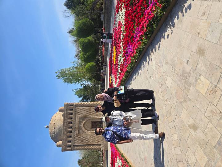 Four travellers pose in front of an ancient brick mausoleum beside colorful flower beds under a clear blue sky.