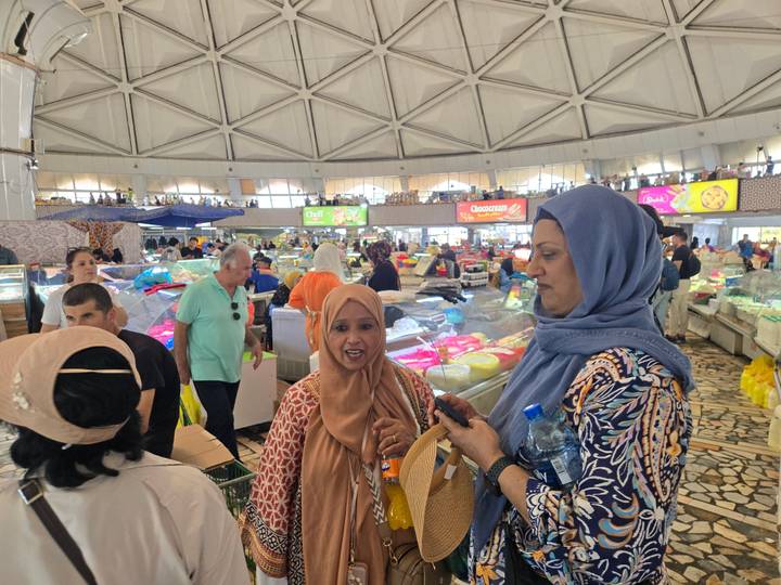 Busy indoor market hall with travelers chatting amid stalls full of colorful produce and cheese wheels.