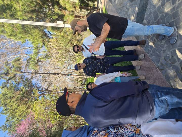 Guide animatedly explains history to a small group standing on a shady park path lined with pines.