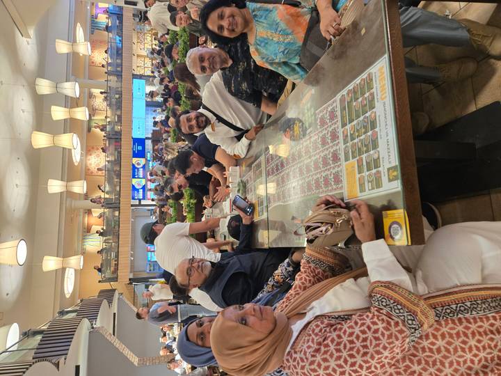 Travellers sit around a long food-court table studying their phones and menus inside a modern mall.