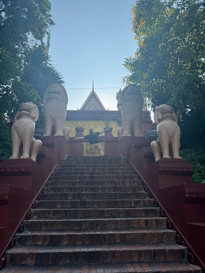 Traveller spreads arms on a flight of temple steps guarded by four lion statues amid thick greenery.
