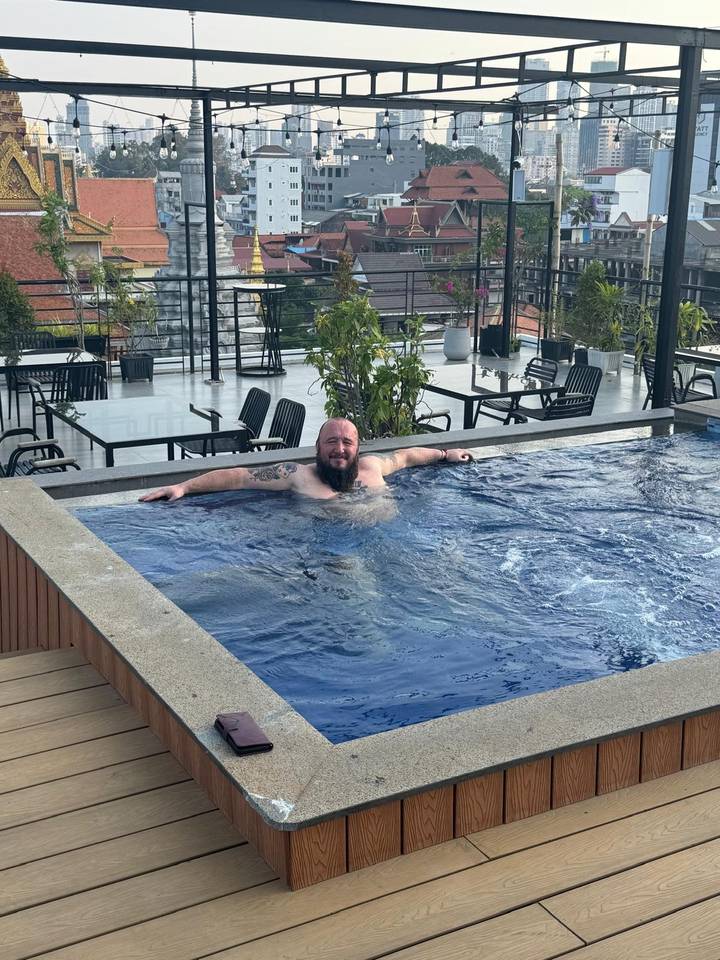 Bearded traveller relaxes alone in a rooftop plunge pool overlooking city buildings.