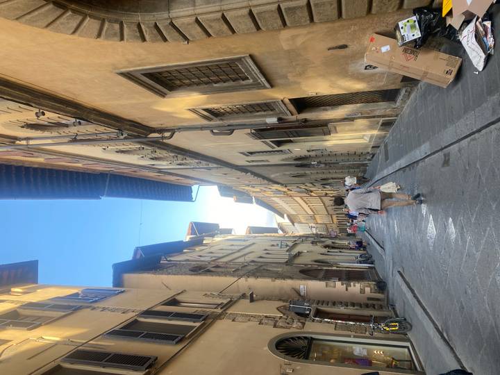Tall yellow-toned buildings frame a pedestrian alley with tourists strolling beneath a clear sky