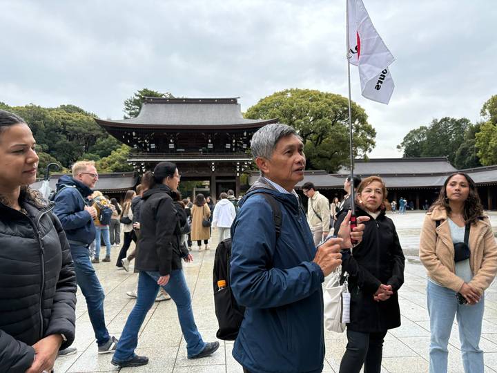 Tour guide holding a flag addresses a group of visitors in the courtyard of a traditional Japanese shrine surrounded by trees.