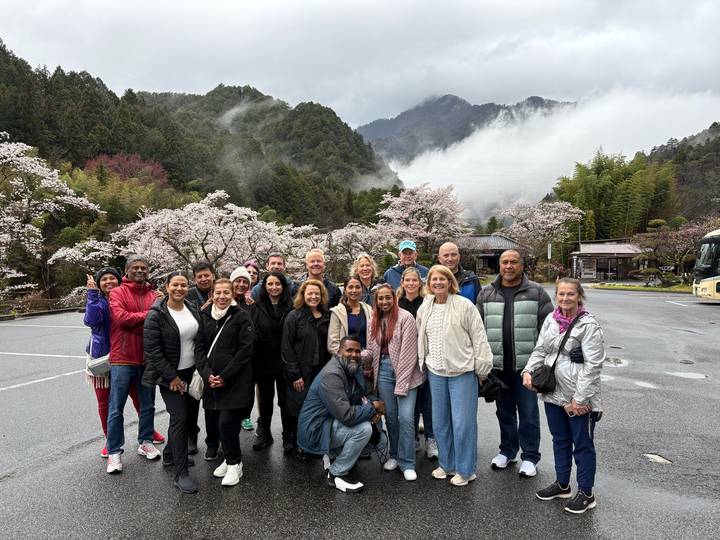 Smiling tour group poses among blooming cherry trees with misty forested mountains rising behind them.