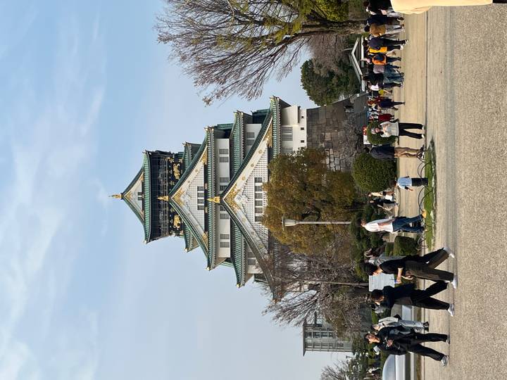 Majestic Osaka Castle with its green and gold roofs rises above a bustling park full of visitors.