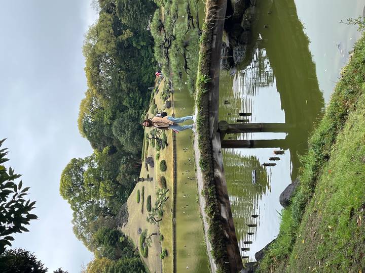 Young woman stands on a moss-covered stone bridge over a tranquil pond in a manicured Japanese garden.