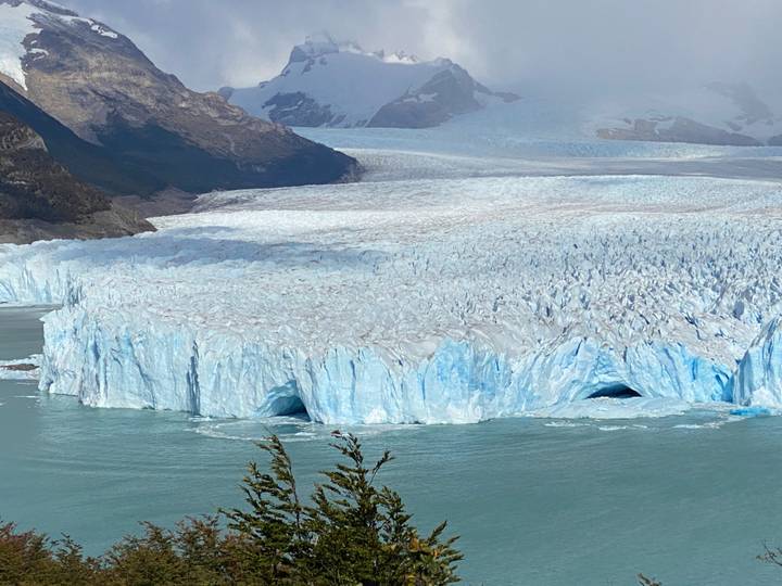 Massive blue wall of Perito Moreno Glacier meets turquoise lake water with distant mountains looming.
