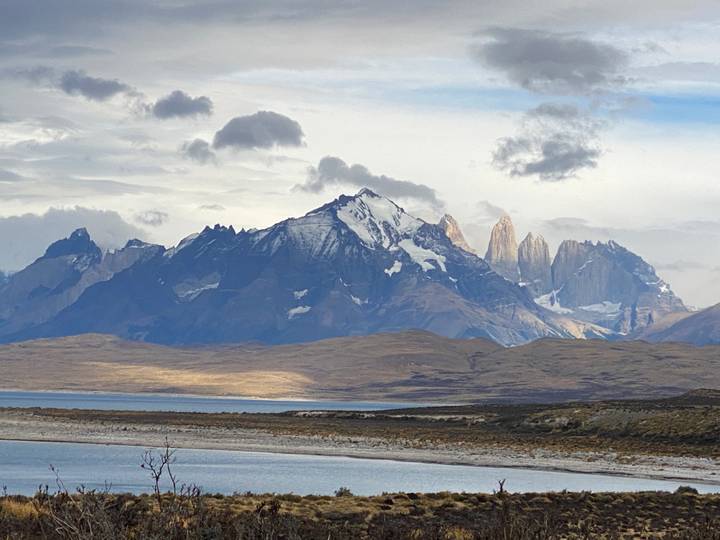 Patagonian plain with a distant turquoise lake and the jagged Torres del Paine massif beneath rolling clouds.
