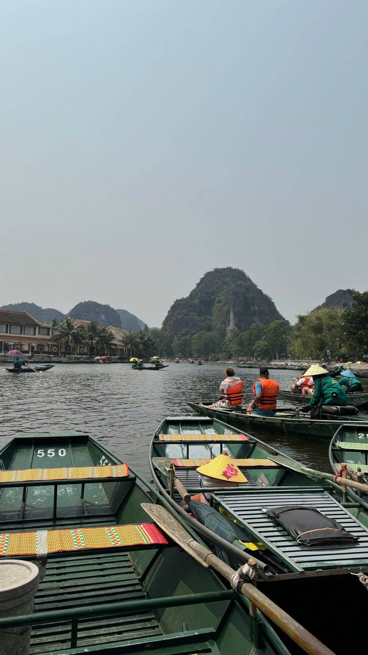 Small wooden boats piloted by locals row tourists along a wide river framed by limestone karst hills.