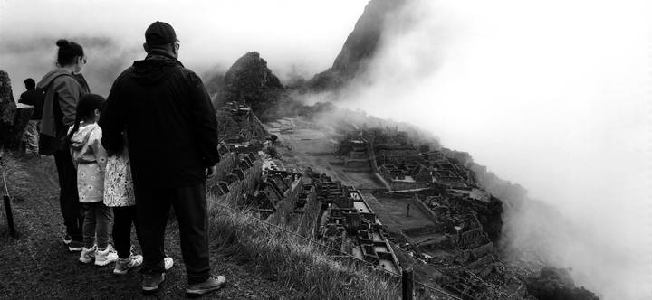 Silhouetted family looks over the mist-covered ruins of Machu Picchu in a dramatic black-and-white scene.