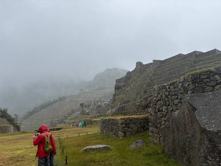 Rain-soaked visitors explore lower terraces of Machu Picchu on a foggy day, stone walls darkened by moisture.