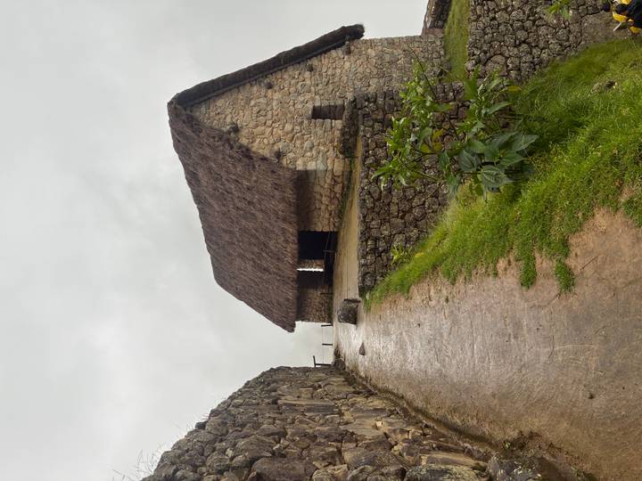 Stone hut with thatched roof sits on a misty path lined by ancient walls at Machu Picchu.