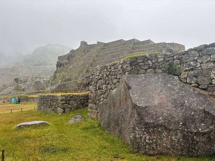Stone terraces and ancient stone walls of Machu Picchu shrouded in mist, with small groups of tourists in colorful ponchos on the grassy foreground.