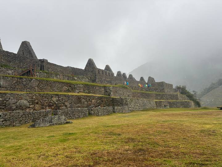 Row of triangular-roofed Incan stone buildings and terraces at Machu Picchu on a cloudy day, with a few visitors walking along.