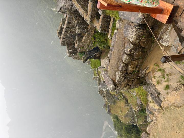 View looking down from a high terrace at Machu Picchu, showing stone walls, steep drop and a misty green valley below.