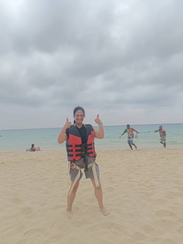 Smiling woman giving thumbs up on sandy beach wearing life vest while staff pull parasail rope behind.