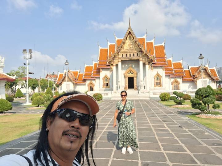 Selfie of two travelers in front of ornate Bangkok temple with orange roofs and manicured garden.