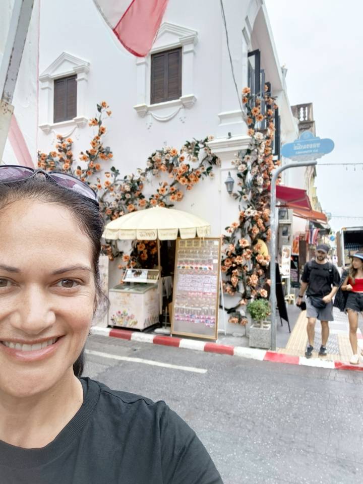 Selfie of a woman on a colorful old-town street decorated with orange flowers and passing pedestrians.