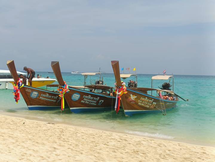Three traditional long-tail boats with colorful garlands rest in shallow turquoise water along a tropical beach.