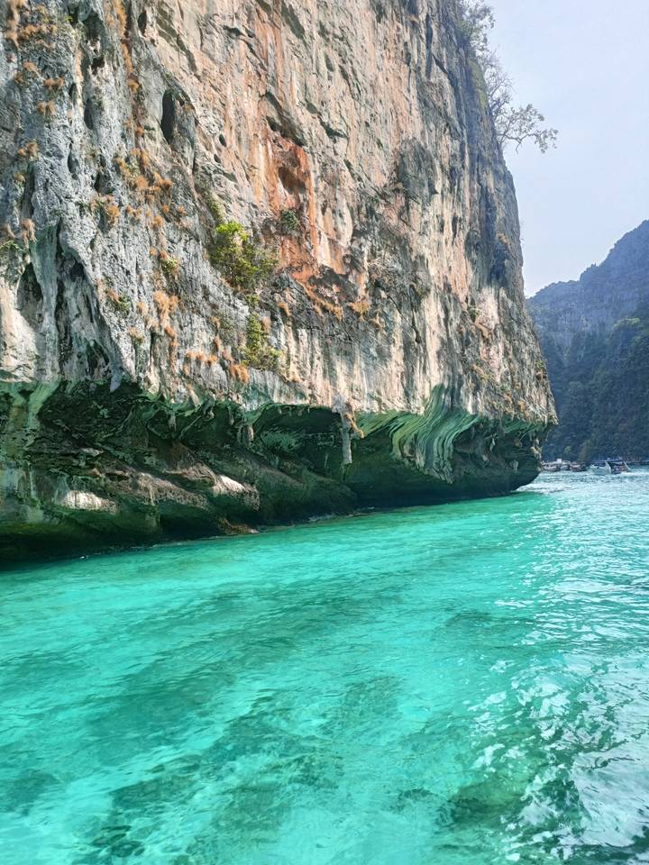 Close-up of a dramatic limestone cliff over bright turquoise sea with boats in the distance.