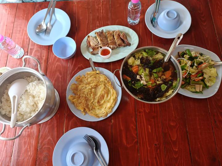 A spread of Thai dishes including rice, omelette, vegetables and grilled meat on a wooden table.