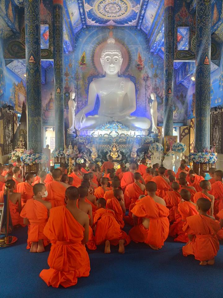 Dozens of novice monks in orange robes sit before a giant white Buddha in a richly decorated hall.