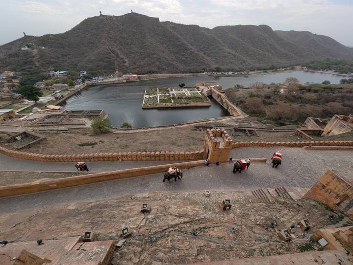 High view over Amber Fort ramparts, elephants with riders climbing the cobbled path beside a lake and gardens