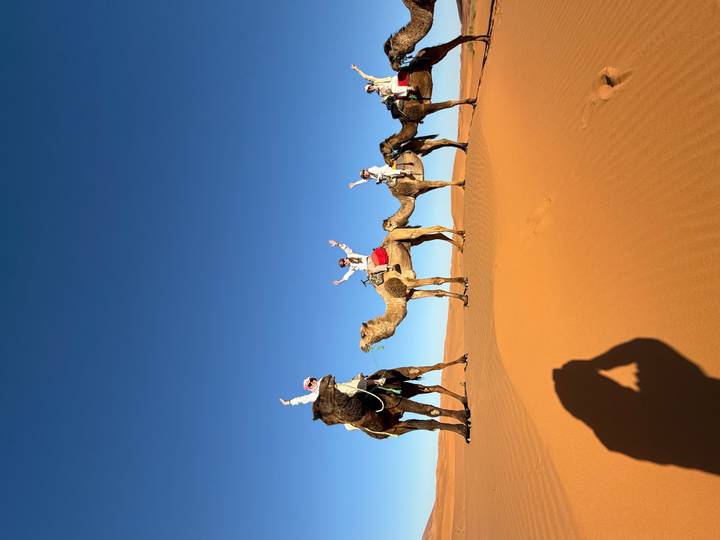 Camel caravan of travellers raising arms while riding across towering orange dunes under a deep blue sky