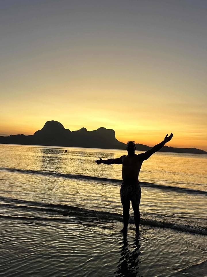 Silhouette of a man with outstretched arms on a beach at vibrant sunset with island backdrop.