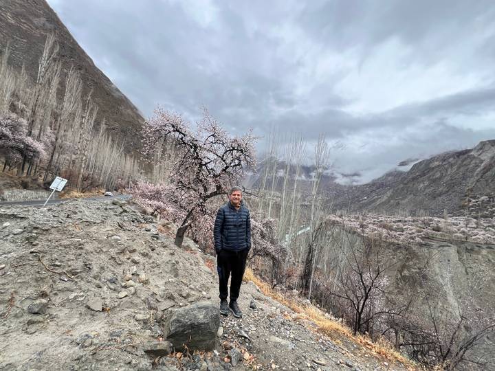 Man standing on rocky roadside next to flowering tree with dramatic cloudy peaks behind