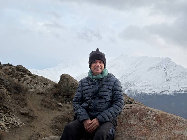 Seated man in winter gear smiles with snowy peaks behind him