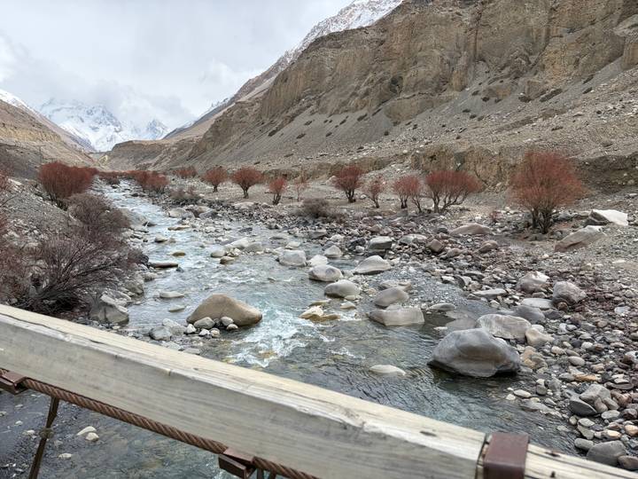 Rocky alpine river flowing through a barren valley lined with small red-leafed trees and distant snowy peaks.