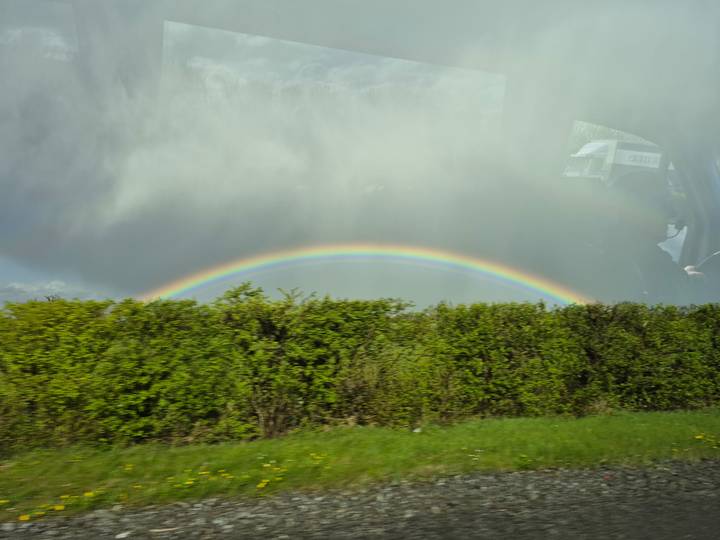Vibrant rainbow arcs above a lush roadside hedge below brooding grey clouds, viewed through a vehicle window.
