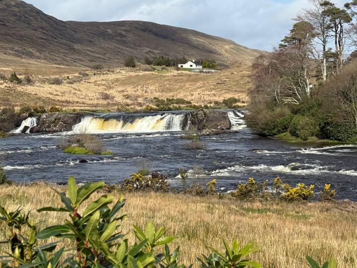 A small waterfall flowing across a rocky river in rural countryside with a lone white cottage on rolling hills behind.