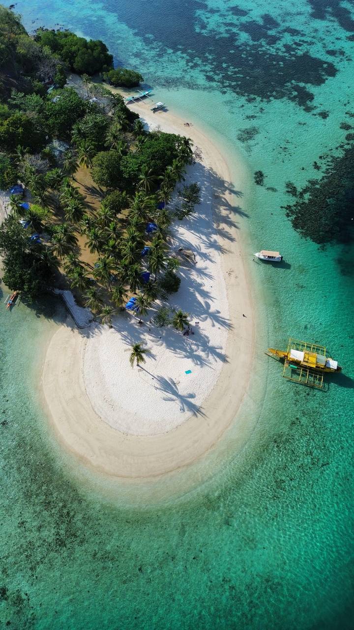 Drone view of a tiny palm-covered island with white sand beach surrounded by clear turquoise water and an anchored yellow boat.