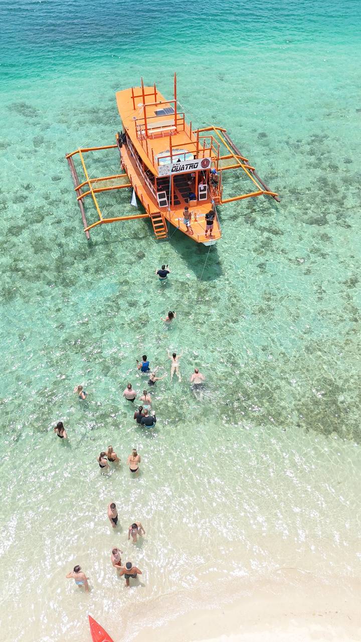 Aerial view of swimmers enjoying crystal-clear shallow lagoon around an orange boat’s ladder.