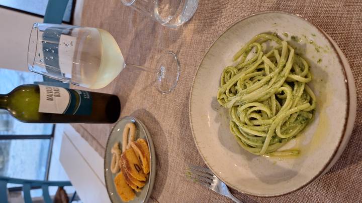 A bowl of pesto linguine with white wine, snacks and a bottle on a rustic table.