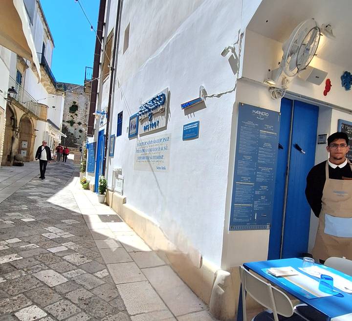 A narrow whitewashed alley with blue accents and restaurant staff welcomes visitors.