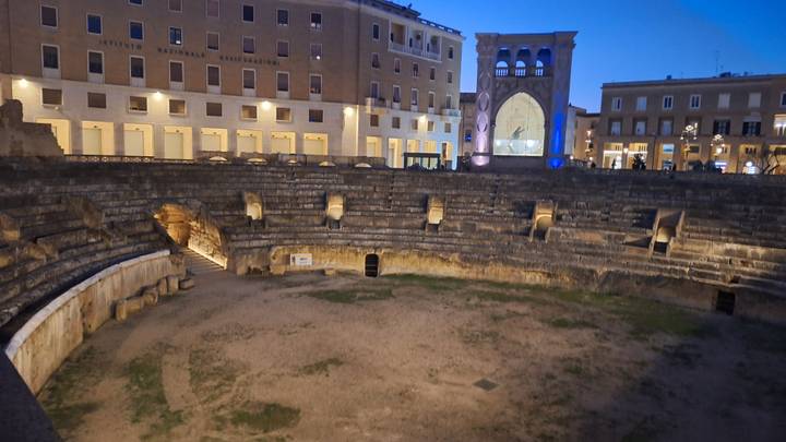 Ancient Roman amphitheatre in Lecce illuminated at dusk, surrounded by historic buildings.