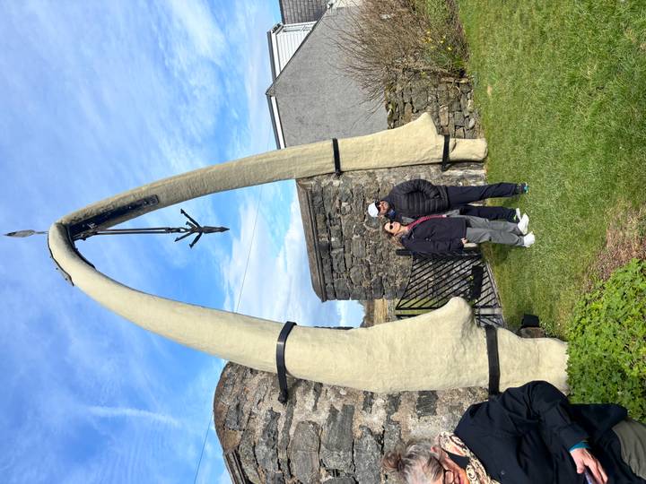 Two travellers pose beneath a giant whale-bone arch outside a stone wall on a bright day