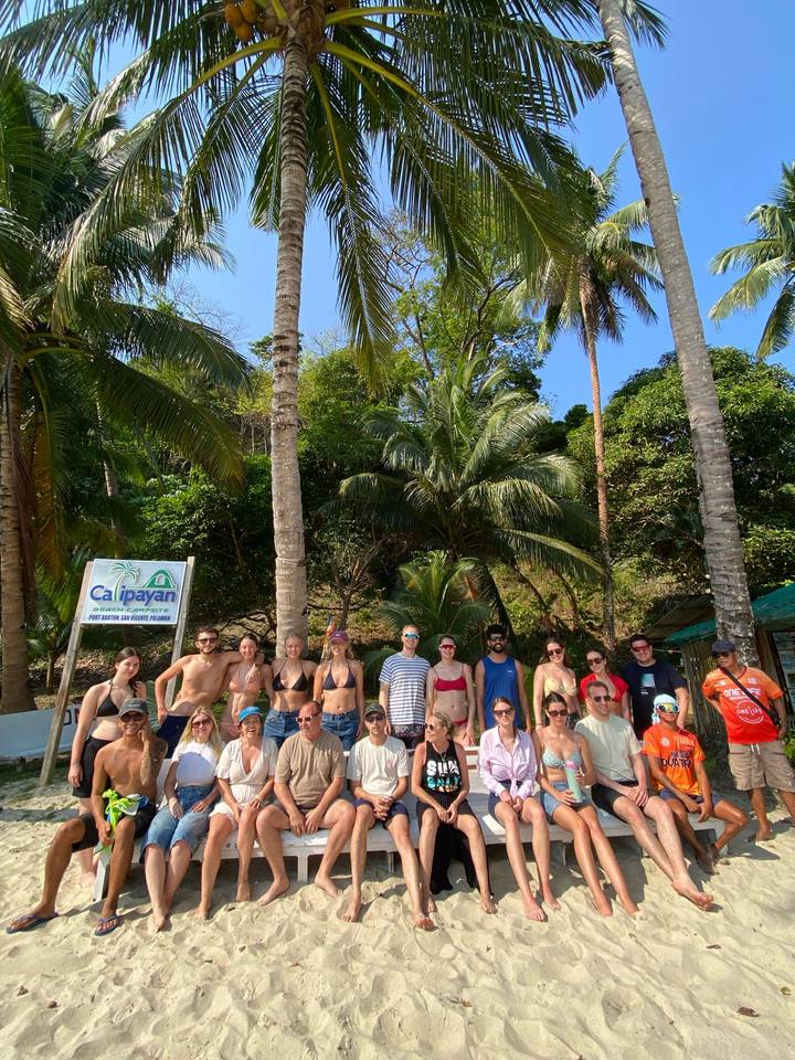 Large tour group posing on a tropical beach backed by palms and lush green hillside.