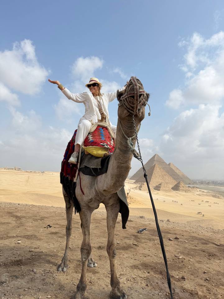 Woman triumphantly riding a camel with iconic pyramids rising from the desert sands behind