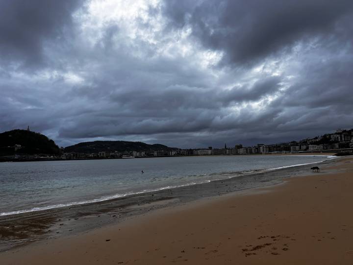 Wide sandy urban beach and bay under dramatic dark clouds with city skyline in background.
