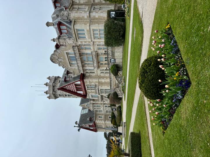 Elegant historic mansion with turrets and red timber detailing surrounded by formal lawns and flower beds.
