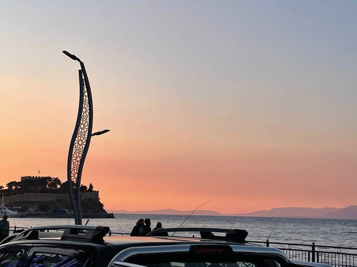Silhouettes of people fishing beside the sea at sunset with an island fortress and modern sculptural streetlight.