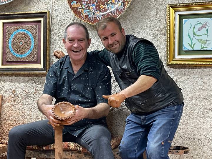 Two smiling men in a pottery workshop displaying a freshly shaped clay bowl, hands covered in clay.