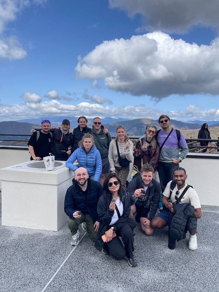Smiling tour group posing at mountain viewpoint with snow-capped Mount Fuji framed by clouds behind.