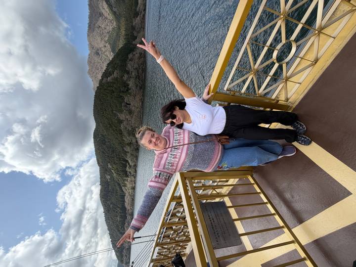 Two happy friends spread their arms on a lakeside ship’s deck with forested hills and water behind.
