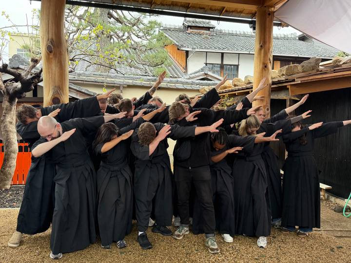 Same robed group striking playful dabbing pose beneath wooden pillars in traditional setting.